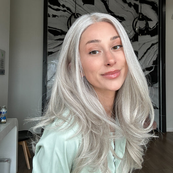 A woman with long, wavy, platinum-blonde hair wearing a pale mint green top, posing in front of a black and white marble wall art piece.