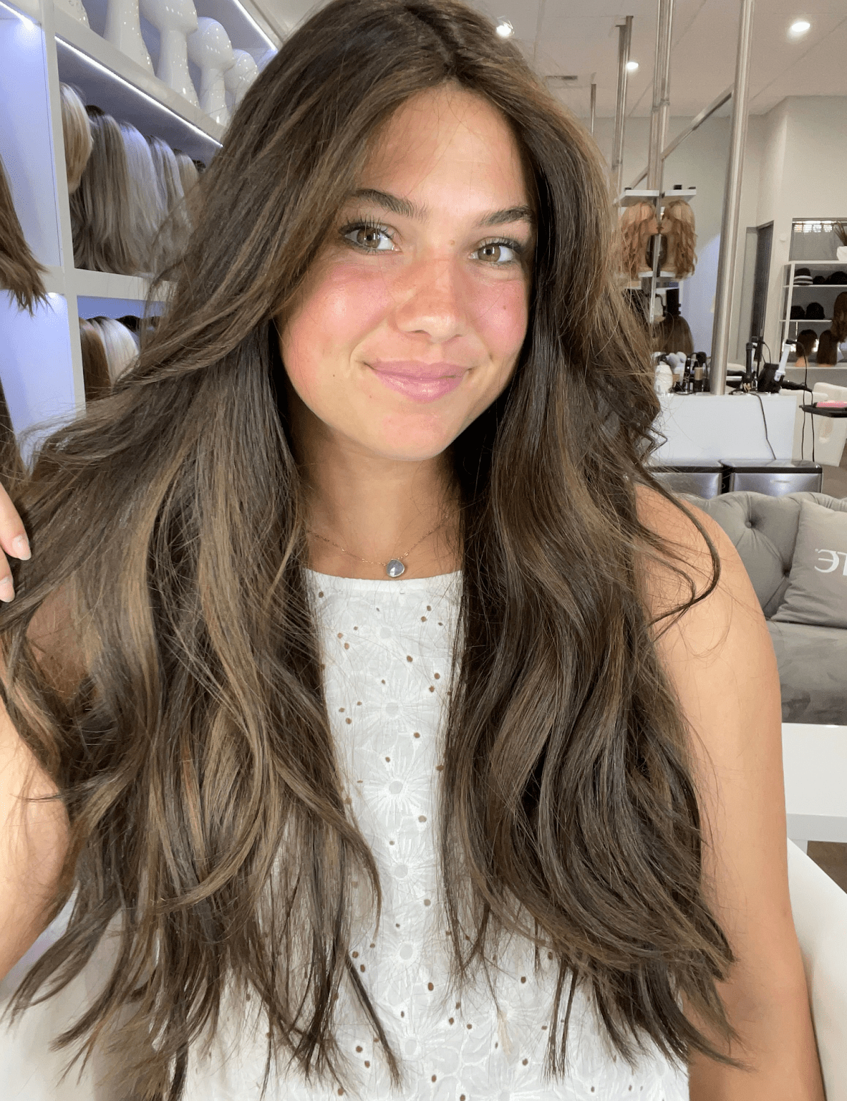 Smiling woman with long, layered brunette waves and soft caramel pieces, in a room lined with wigs on mannequin heads