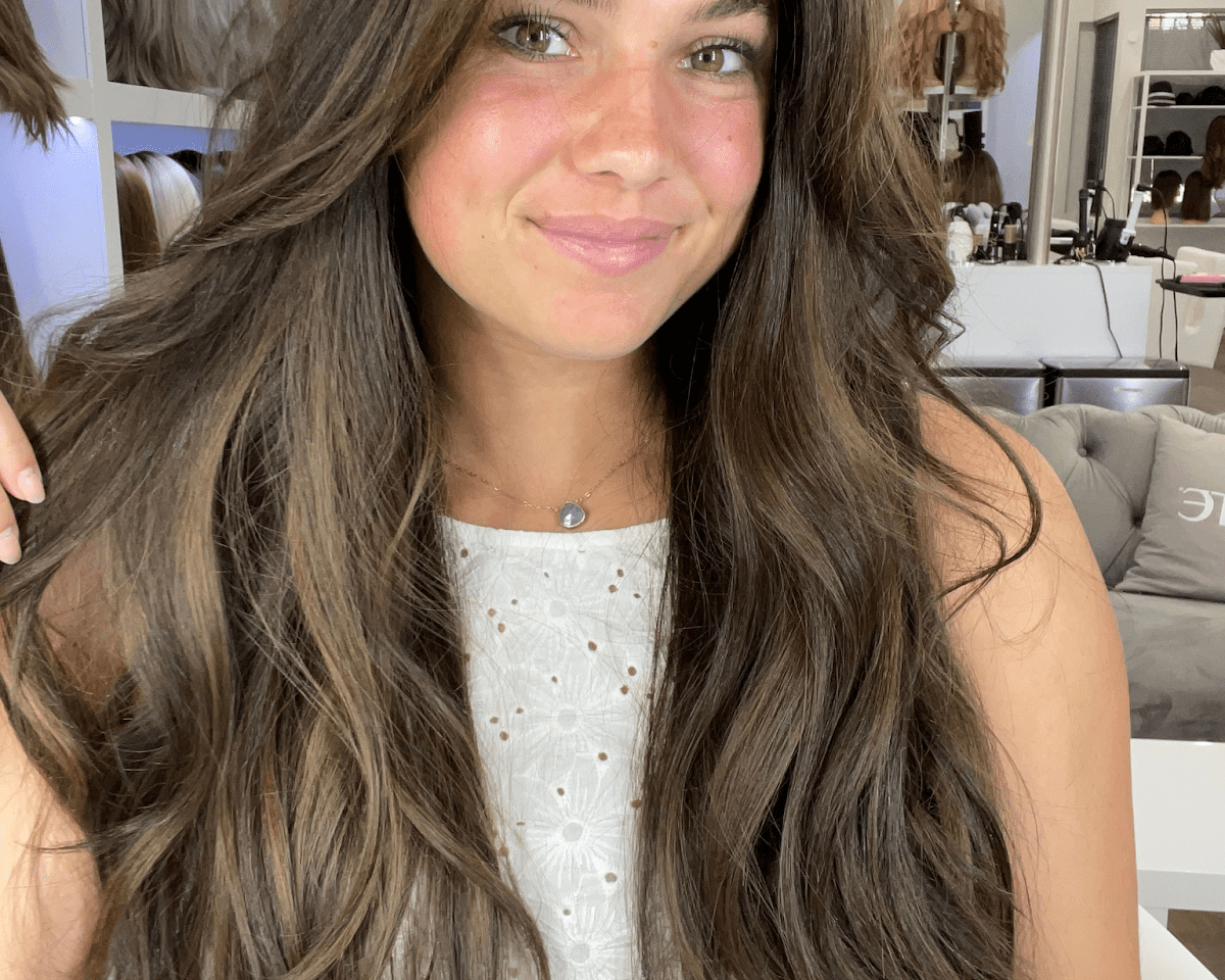 Smiling woman with long, layered brunette waves and soft caramel pieces, in a room lined with wigs on mannequin heads