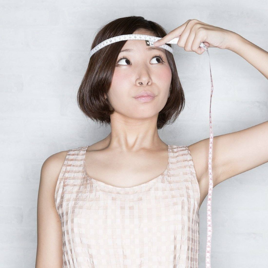 Woman measuring her head with a tape measure against a plain background